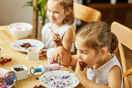 two beautiful sisters in white dresses decorate and eat delicious cupcakes at a wooden tableの写真素材