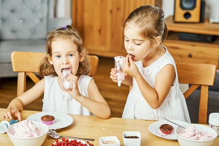 two beautiful sisters in white dresses decorate and eat delicious cupcakes at a wooden tableの写真素材