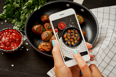 woman's hands are holding the phone and taking a picture of delicious meatballs on a wooden tableの写真素材