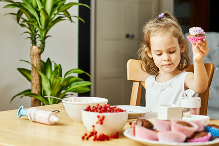 pretty girl in a white dress decorates and eats a delicious cupcakes at the wooden tableの写真素材