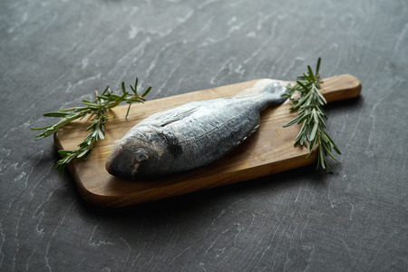one fresh raw dorada fish with rosemary, pepper and salt on a wooden board and a black table