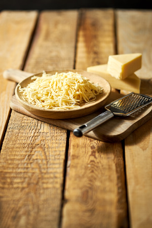 Shredded yellow cheese on wooden plate with cheese grater on kitchen tableの写真素材