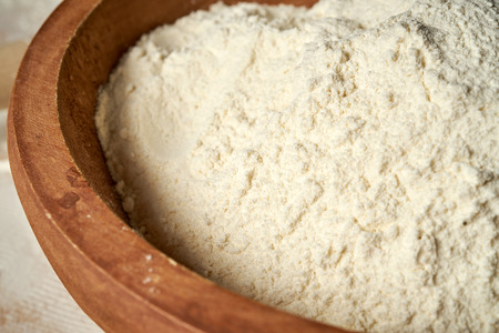 Closeup of flour in wooden bowl on white wooden table.