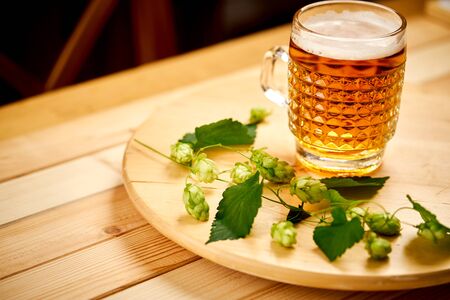 Large pint of beer with foam and sprigs of hops on wooden table