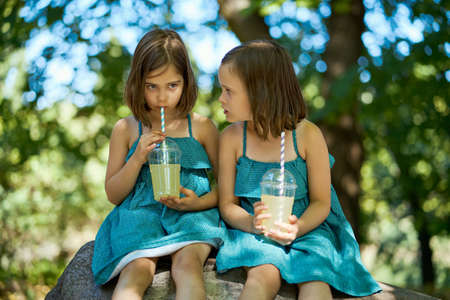 two cute little girls in dresses sit on a stone and drink lemonadeの写真素材