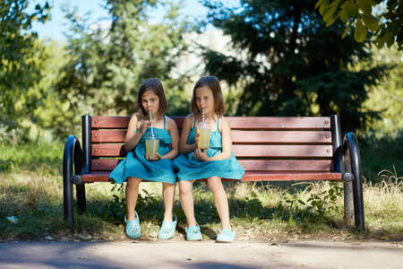 two cute little girls in dresses sit on a bench and drink lemonade in the summerの写真素材