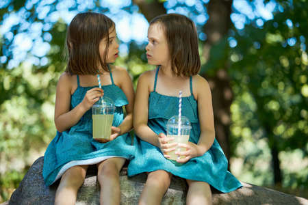 two cute little girls in dresses sit on a stone and drink lemonadeの写真素材