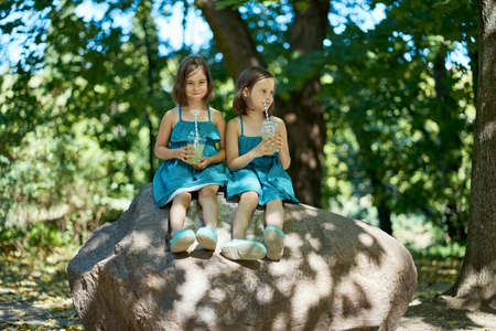 two cute little girls in dresses sit on a stone and drink lemonadeの写真素材