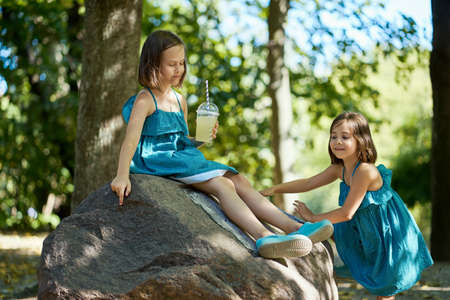 two cute little girls in dresses sit on a stone and drink lemonadeの写真素材