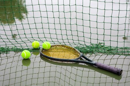 Tennis balls and racket on wet tennis court after raining. Racket and tennis balls laying beside a...