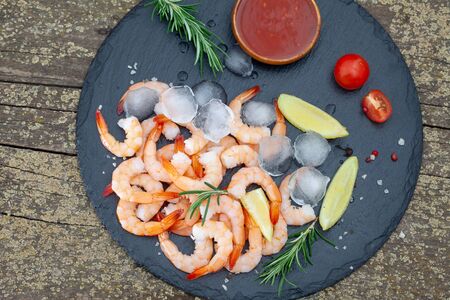 Fresh shrimps with lemon, rosemary and sauce on black plate. Shrimp tailes on ice, top view.の写真素材
