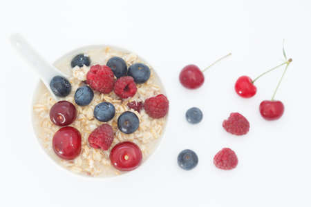 Rolled oats with blueberries, raspberries and sour cherries on white background. Healthy breakfast cereal oat flakes in white bowl, top view.の写真素材