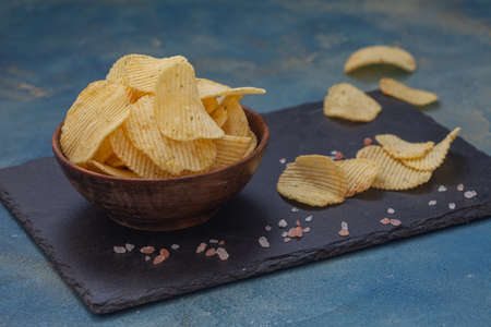 Crispy potato chips in a wooden bowl on blue background. Salty crisps scattered around.の写真素材