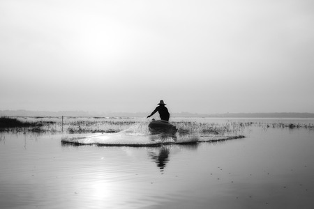 Black and white of fishermen casting for catching the fish on the wooden boat at the lake in the morning. Thailand lifestyle.の写真素材