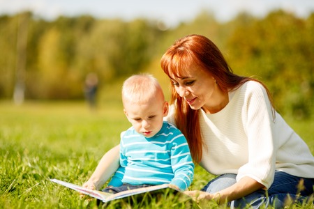 Picture of beautiful woman with son sitting down on green grass field and read fairytale in parkの写真素材