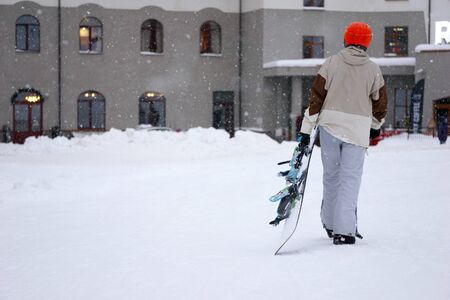 Taired man snowboarder wearing orange helmet, grey jacket, black and blue glovesa and grey pants with snowboard in one hand coming back in hotelの写真素材