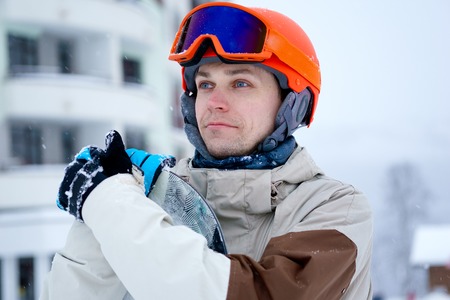Man snowboarder wearing orange helmet, grey jacket, black and blue gloves standing with snowboardの写真素材