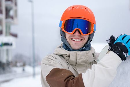 Man snowboarder wearing orange helmet, grey jacket, black and blue gloves standing with snowboardの写真素材