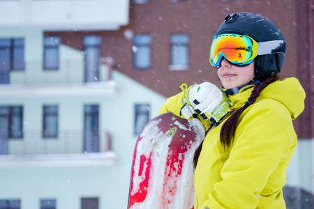 Female snowboarder wearing black helmet, yellow jacket, white and green gloves standing with snowboardの写真素材