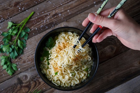 Top view of hands with chopsticks holding chinese noodles with fresh peas and chopped green onion on wooden table.の写真素材