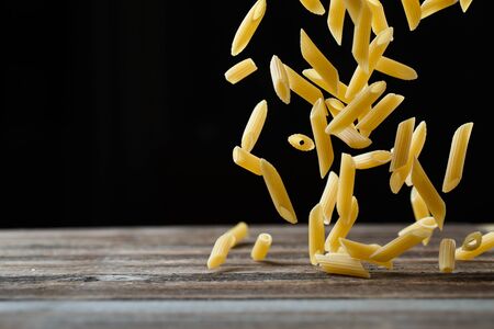 Falling penne pasta. Flying yellow raw macaroni over black background and rebound from wooden table....