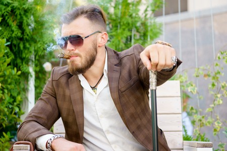 Caucasian stylish man with sunglasses sitting with cane and smoking cigarの写真素材