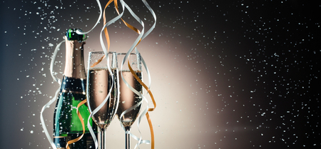 Green bottle of champagne, wine glasses with silver and golden ribbons close-up. Festive composition on a dark background with backlightの写真素材
