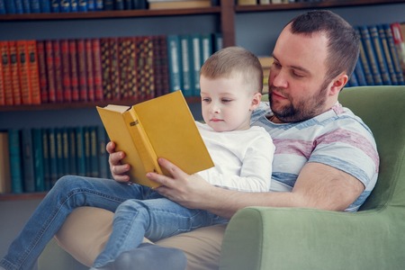 Photo of dad reading to son book sitting in chairの写真素材