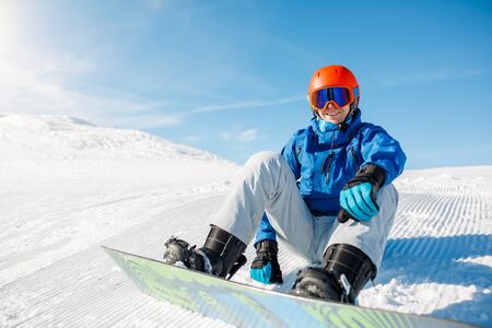 Photo of sports man wearing blue jacket, helmet with snowboard sitting on snowy slopeの写真素材
