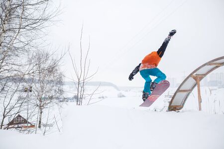Photo from back of young athlete skating on snowboard with springboard against background of treesの写真素材