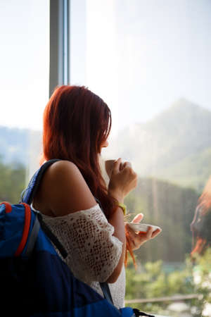 Long-haired woman with cup of tea and backpack near window