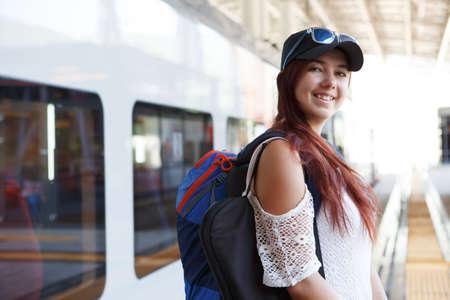 Photo of smiling woman tourist in cap and sunglasses with backpack near trainの写真素材