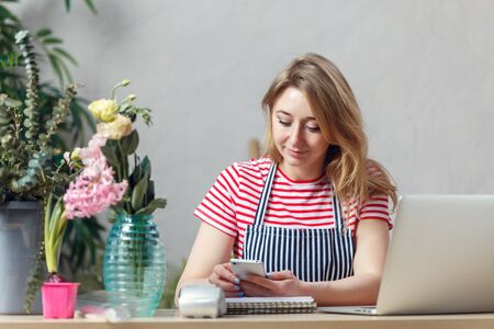 Photo of florist girl with phone in hands sitting at table with computer