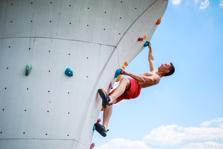 Photo from bottom of strong athletic guy exercising on wall for climbing against blue sky with cloudsの写真素材