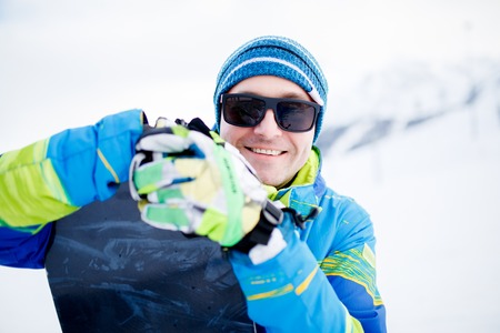 Portrait of happy man in sunglasses with snowboard in hands on background of snowy mountainsの写真素材