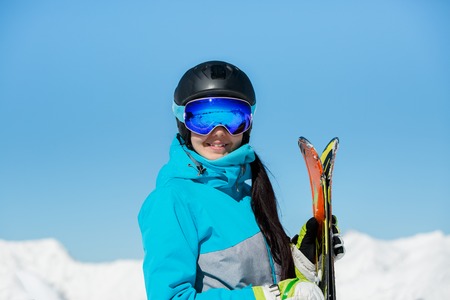 Photo of young smiling female athlete in helmet with skis in hand against blue sky and snowy hillの写真素材