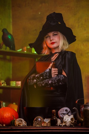 Witch in black hat with spell book at table with pot, pumpkin