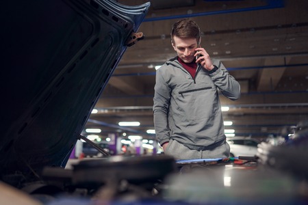 Young man talking on phone standing by car with open hood in underground parking