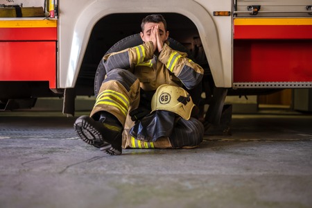 Picture of tired fireman sitting on floor near red fire truckの写真素材