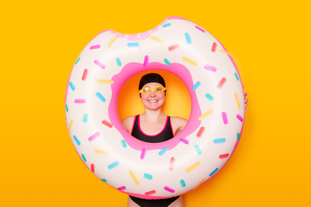 Photo of girl in swimming goggles, swimsuit with donut life buoy on empty orange background