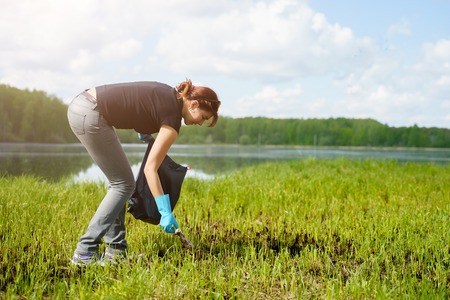 Photo of side of brunette in rubber gloves picking up garbage in bag on bank of river.