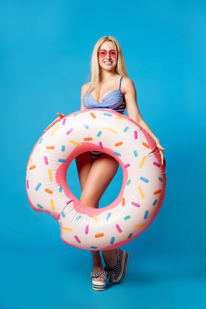 Full-length photo of young woman in bathing suit with inflatable donut on empty blue backgroundの写真素材