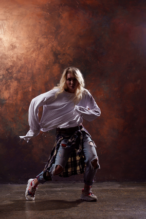 Photo of young dancing blonde girl in ripped jeans and sneakers on brown backgroundの写真素材