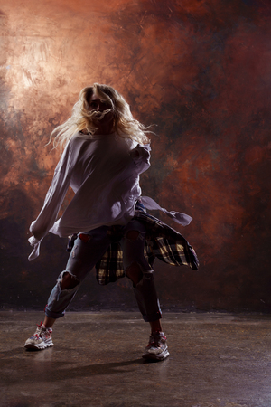 Photo of young blonde dancer in torn jeans and sneakers on dark brown backgroundの写真素材