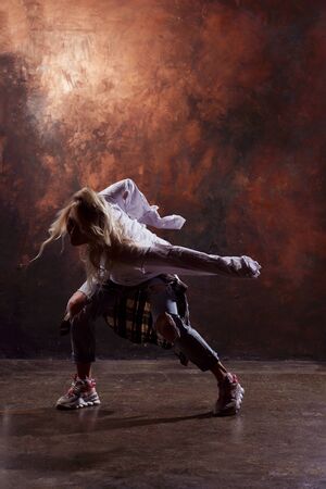 Photo of slim blonde dancer in torn jeans and sneakers on dark brown backgroundの写真素材