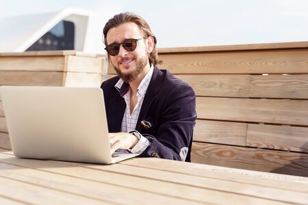 Photo of cheerful man in sunglasses sitting with laptop on wooden bench on spring