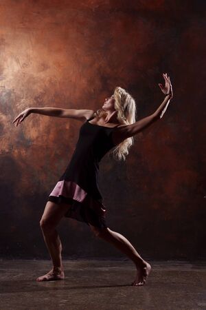 Photo of young dancing blonde looking back in black dress on brown backgroundの写真素材