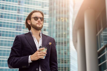 Photo of brunet man with beard looking to side with glass in hand in city against background of tall buildingsの写真素材