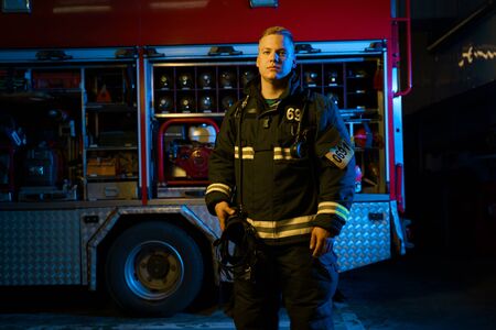 Photo of firefighter with helmet in his hand looking into camera while standing near fire truck