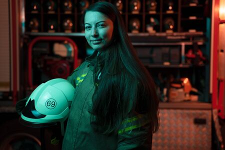 Photo of woman firefighter with long hair with helmet in her hands at fire station.